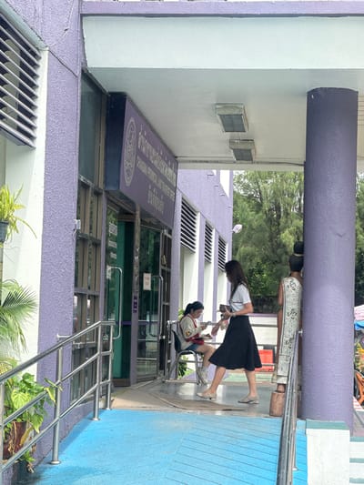The image depicts the exterior of the Chiang Mai Provincial Land Transport Office in Mae Hia Town Municipality, Thailand. A purple building with white trim and horizontal louvers is visible. In front of the building, a ramp with a blue surface and a silver railing leads to the entrance. Two women are standing near the entrance. One woman, wearing a white t-shirt and a black skirt, is holding a book and looking to her left. Another woman, wearing a white sweater and denim shorts, is seated on a chair to the left, looking down at her phone. A decorative statue of a person wearing traditional clothing stands to the right of the entrance, partially obscured by a purple pillar. Lush green trees are visible in the background.