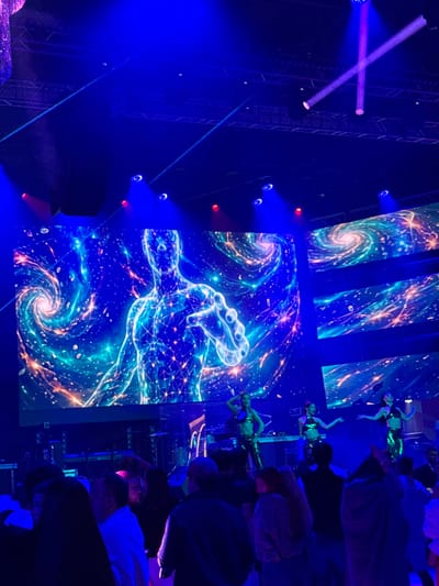 The image depicts a vibrant concert or performance event illuminated by an abundance of blue and purple lights. In the background, a large screen displays a cosmic-themed visual featuring a glowing human-like figure outlined in blue circuitry, surrounded by swirling galaxies and stars. On a stage in front of the screen, three performers in sparkly outfits are dancing. Below the stage, a crowd of people is visible, some with their backs to the camera, appearing to be enjoying the show. The overall atmosphere suggests a lively and energetic entertainment venue, possibly a nightclub or concert hall. The mention of Akhalubani, Georgia, suggests this event is taking place in that city, though the visual details themselves do not provide specific geographic clues.