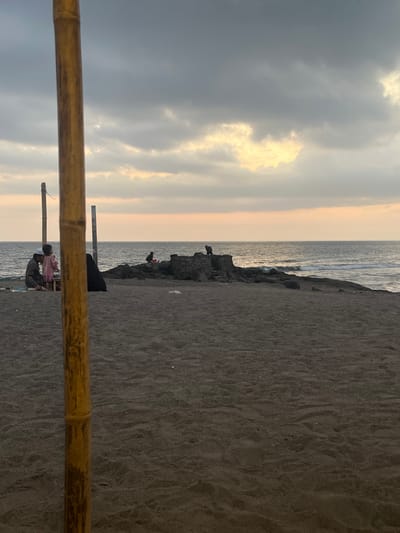 The image depicts a beach scene in Desa Batu Layar, Indonesia, at dusk or dawn. The sky is predominantly overcast with grey clouds, exhibiting a horizontal band of pale orange-yellow light along the horizon over the ocean. The ground consists of dark grey sand. A prominent yellow-brown bamboo pole extends vertically along the left edge of the frame, partially obscuring the view. To the left, behind this pole, a person in dark clothing is partially visible. Further left, an adult, dressed in a light-colored top and dark pants, is seated on the sand next to a standing child in a pink garment. Two vertical wooden posts are positioned behind this human group. In the midground-right, a dark rocky outcrop extends into the water. Two dark-furred monkeys are on these rocks: one crouches on a lower section, and another stands on a raised, square rock formation. A third human figure is also present on the rocky outcrop, situated to the left of the monkeys. The ocean surface exhibits small waves near the shoreline.