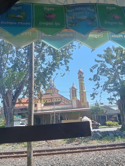 The image captures a view of a mosque in Sruni, Indonesia, framed by a canopy displaying advertisements for "Kelapa Muda" (young coconut) and "Degan Ijo" (green coconut). The mosque has a red-tiled roof and a distinctive yellow and green minaret topped with a patterned dome. In the foreground, railway tracks run parallel to a road where a truck and a car are visible. The sky is clear and blue, with lush green trees flanking the scene. The overall impression is of a typical day in a tropical Indonesian town.