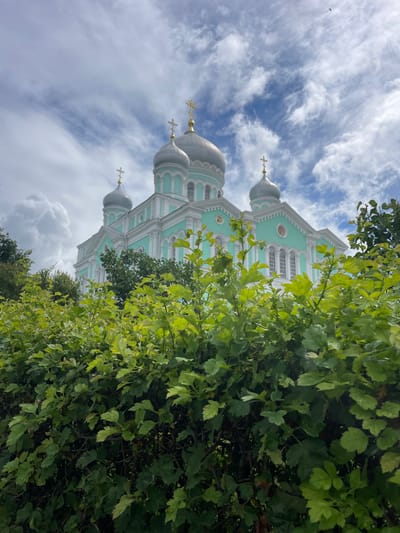 The image shows a large, light teal and white church with several silver domes topped with gold crosses. The church is viewed from a low angle, with lush green foliage in the foreground obscuring the lower portion of the building. The sky is partly cloudy with wispy white clouds against a blue background. The church is the Seraphim-Diveyevo Monastery, located in Diveyevo, Russia. No people are visible in the image.