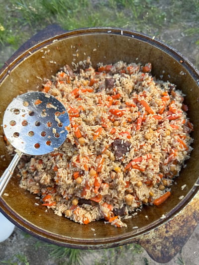 A close-up, overhead shot displays a large, well-seasoned kazan filled with pilaf. The pilaf consists of rice, diced carrots, chickpeas, and chunks of dark meat, possibly beef or lamb. A perforated metal ladle rests partially in the pilaf, its handle extending towards the bottom left of the frame. The kazan itself is dark, metallic, and shows signs of use, with a thick, textured rim. The background consists of blurred green grass and earth, suggesting an outdoor setting, likely in a rural area or park. The scene is set in Bolshiye Dvory, Russia, implying a traditional outdoor cooking or communal meal.