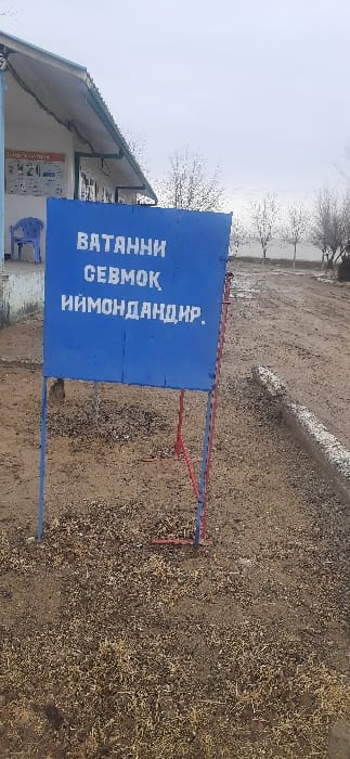 A blue sign with white lettering is displayed on a red and blue metal stand. The text on the sign is vertical. The sign is positioned in a landscape of dry soil and sparse vegetation. A building with a slanted green roof is partially visible on the left. In the background, barren trees and a body of water can be seen under a cloudy sky.