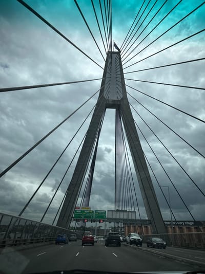 A low-angle shot showcases the Anzac Bridge in Sydney, Australia. The bridge's tower dominates the frame, with numerous suspension cables radiating outwards against a backdrop of a cloudy sky tinged with blue. A roadway runs alongside the bridge structure, carrying vehicular traffic. Signs and structures associated with the road are visible on the right, completing the scene.
