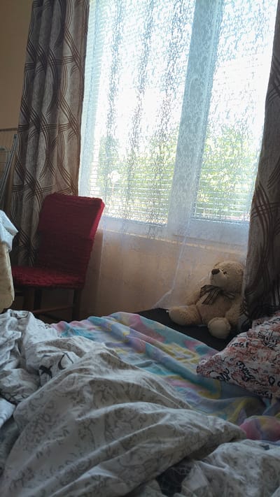 A dimly lit bedroom scene is captured with the focus on a rumpled bed with a colorful comforter and patterned sheets. To the left of the bed is a red chair covered in a textured fabric. A teddy bear with a plaid bow sits on the edge of the bed. Behind the bed, a window is covered by sheer lace curtains and blinds, with a view of green trees outside. The overall impression is one of a cozy and lived-in space. The image is a photograph taken inside a room, possibly in a residential setting in Han Krum, Bulgaria.