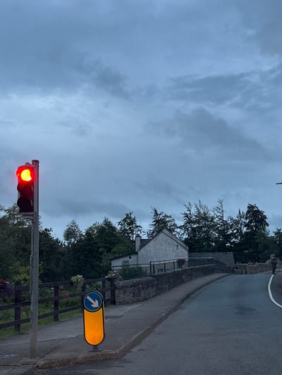 The image shows a traffic light displaying a red signal, located at an intersection in Rathvilly, Ireland. The traffic light is positioned on the left side of the road, with a pedestrian crossing signal illuminated yellow and displaying a blue circular sign with a white downward-pointing arrow. The road curves to the right, leading over a stone bridge. On the bridge, there is a low stone wall with a railing and a building with a light-colored facade. Trees and foliage line the sides of the road and the bridge. The sky is overcast with dark clouds. Two individuals are visible in the distance: one standing on the right side of the road near the bridge, and another, partially obscured, further down the road.