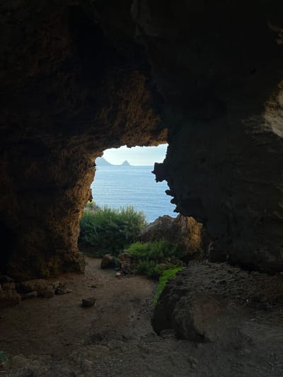 The image displays a forensic view from within a natural rock cave in Sperone, Italy, looking outwards towards the sea. The cave interior, occupying the majority of the frame, is largely shadowed, revealing rough, textured rock walls and ceiling. The ground inside the cave is composed of dark, uneven dirt with several scattered rocks. A substantial, irregularly shaped opening in the rock face frames the exterior environment.
Through this opening, a calm, deep blue body of water is visible under a bright sky, extending to a distant horizon. On the horizon, two distinct, light-colored mountainous landmasses or islands are silhouetted against the sky. Immediately outside and below the cave opening, a vibrant cluster of green bushes and leafy vegetation thrives on an elevated section of land. No human figures, animals, or man-made objects are present in the scene. The lighting emphasizes the stark contrast between the darkened, shadowed cave interior and the brightly illuminated exterior landscape.