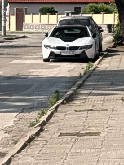 The image displays a daytime street scene in Saedinenie, Bulgaria. In the middle ground, a white BMW i8 coupe is parked facing left, with its front license plate, "CB 9336 OB," clearly visible. Partially obscured behind the BMW i8 is a dark-colored vehicle. The vehicles are positioned on an asphalt road featuring faded white road markings, consistent with a pedestrian crossing, situated to their left. A concrete curb separates the asphalt road from a light-colored paved sidewalk on the right side of the frame. Small green plants are growing in the cracks of both the curb and the sidewalk pavers. In the background, across the street, a building with a red-tiled roof and yellow exterior walls is visible, featuring a dark brown wooden door. A low, light-colored decorative fence or wall stands in front of this building. Green foliage, consistent with trees or bushes, is present above the background structures. Distinct shadows cast by the vehicles and the curb indicate bright sunlight. No individuals are visible in the scene.