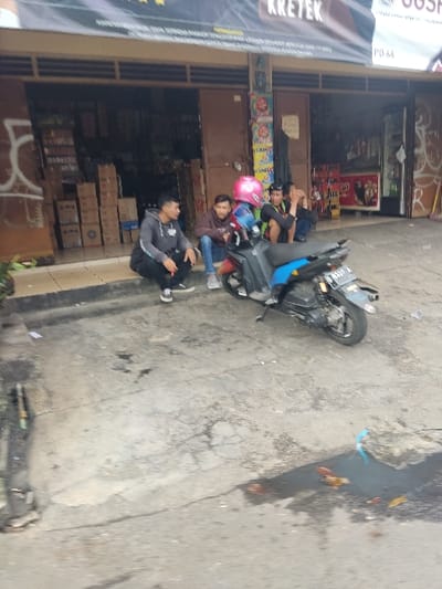 The image shows a scene outside a shop in Kecamatan Cimahi Selatan, Indonesia, located at the coordinates -6.903 latitude and 107.564 longitude. The shop has a brown awning with the word "KRETEK" written on it in white lettering. There are several boxes stacked in front of the shop, and the shop is open, with a man standing on the sidewalk in front of it. There are three men sitting on the sidewalk in front of the shop. One of the men is wearing a pink helmet, another is wearing a blue shirt and a third is wearing a green shirt. There is a blue and silver motorcycle parked in front of the men. The street is paved and there are some plants in the foreground.