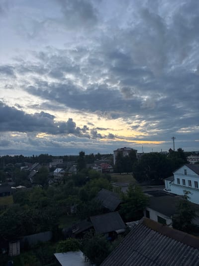 The image depicts an aerial view of a village or small town with a dramatic, cloudy sky at dusk. The sky is filled with a mixture of dark gray, heavy clouds and lighter, thinner clouds that allow patches of pale yellow and orange light to peek through, suggesting the setting sun. Below the sky, a dense arrangement of residential buildings, mostly houses with sloped roofs, is visible, interspersed with numerous trees and greenery. Some of the buildings have visible details like windows and chimneys. In the background, taller structures, including what appears to be a communication tower and a multi-story building, can be seen, indicating a more developed area on the horizon. The overall impression is one of a quiet, rural or semi-urban environment under a moody sky. The location specified, Пратасевіцкі сельскі Савет, Belarus, suggests a Belarusian setting.