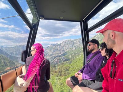 Inside a cable car, four individuals are seated, observing the landscape. The person furthest left has pink braided hair, wearing sunglasses, a dark top, and shorts, with a light-colored bag slung over their shoulder. To the right, a man with a beard wears a black cap, sunglasses, and a purple hoodie. Beside him, a woman with dark curly hair dons sunglasses and a dark jacket. The man on the right wears a red cap and shirt.

Outside the cable car, the vista reveals mountains and a bay. The location is near Mirac, Montenegro, as evidenced by the bay and mountainous landscape. The sky features a mix of blue and white clouds. The interior of the cable car is primarily dark, with a camera visible on the ceiling. The car's seating is upholstered in a light brown material.