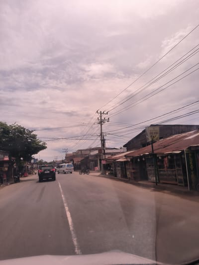 This image captures a street scene in Paya Tringgadeng, Indonesia, viewed from inside a vehicle. The paved road, marked with a faded white line, stretches into the distance, flanked by buildings on both sides.

On the right, a series of humble, single-story shops line the street. These structures feature wooden facades and distinctively rusty, corrugated metal roofs. Goods are visible through display cases in front of the shops. A prominent sign on one building reads "KAK AINA," accompanied by an illustration of a motorcycle and additional text. Further down, a partial sign appears to say "BUKATI." Overhead, numerous utility poles with a dense web of electrical and communication wires crisscross the sky. A person is visible riding a motorcycle on this side of the road.

On the left, a large, leafy tree partially obscures other structures. A sign showing "IMA" and "15.000" (likely a price in Indonesian Rupiah) is partially visible closer to the foreground. Another motorcycle can be seen further down the road on this side.

In the mid-ground, a black SUV, likely a Toyota Innova, is seen from behind, bearing the Indonesian license plate "BL 2696 JAH." Ahead of it, a white van proceeds down the road.

The sky is overcast with light, scattered clouds, indicating diffused daylight, possibly late morning or afternoon. The scene depicts a typical semi-urban or rural thoroughfare, showing everyday life and commerce. Reflections from the car's windshield are visible at the very bottom of the image.