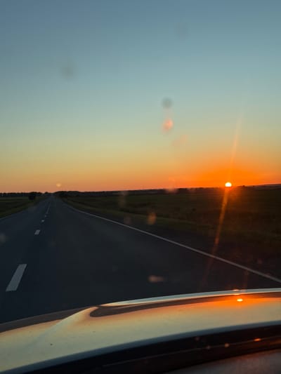 The image shows a road stretching into the distance under a sky with a sunset. The sun is visible as a bright orange orb on the horizon, casting a warm glow across the sky and the landscape. The road has white dashed lines marking the lanes and appears to be made of asphalt. There are fields on either side of the road, with some trees visible in the distance. The foreground shows a portion of a vehicle, possibly a car, with its hood reflecting the colors of the sunset. There are a few blurry circular marks in the sky, which could be dust or water droplets on the lens. The scene suggests a journey on a rural road during a beautiful sunset. Morozovka, Russia is a village located in the Belgorod Oblast.