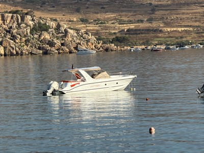 The image captures a section of the coast in St. Paul's Bay, Malta, characterized by a rugged, rocky shoreline with dry, terraced hills in the background. The foreground is dominated by a calm, blue body of water where several boats are anchored. The most prominent boat in the mid-ground is a white motor yacht with orange accents and the name "CALIONBOATS" inscribed on its side. It has a canvas canopy over a portion of the deck and two outboard motors at the stern, one of which is covered. To the left of this yacht, a smaller white boat is partially visible. Further in the background, a line of various other vessels, including sailboats and motorboats, are moored along the coast. Two buoys are visible floating in the water: a small, cylindrical red one and a larger, round red and white one. The overall impression is one of a peaceful, sunny day in a Mediterranean coastal area.