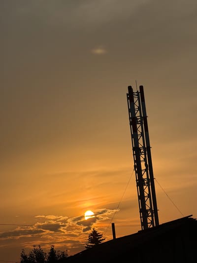 This media file depicts an outdoor scene in Vikulovo, Russia, dominated by a dramatic sunset. The image is oriented vertically, showing the sky with vibrant hues of orange and yellow, indicative of the late afternoon or early evening. Wispy clouds are illuminated by the setting sun, which is partially obscured by them, creating a soft, diffused glow.
In the foreground and mid-ground, industrial elements are silhouetted against the bright sky. A large, metallic structure, possibly part of a heating or cooling system, extends horizontally across the frame. It appears to be made of parallel pipes supported by a metal framework. The structure is painted with alternating red and white bands for visibility. To the right, the dark silhouette of a building, likely an industrial facility or warehouse, rises vertically, further emphasizing the industrial setting. Some sparse vegetation, like trees or bushes, is also visible in silhouette along the right edge of the image.
The overall mood is serene, with the natural beauty of the sunset juxtaposed against man-made structures. The lighting suggests a clear or partly cloudy day transitioning into evening, with the sun creating a warm and atmospheric backdrop. There is no visible text in the image.