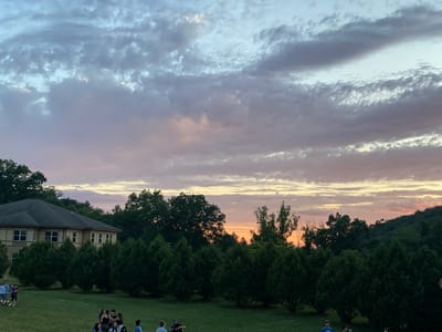 The image depicts a dusky sky with scattered clouds, transitioning from pale blue to hues of pink and orange near the horizon. Below the sky, a row of dense, dark green evergreen trees runs horizontally across the mid-ground, creating a natural barrier. Behind the evergreens, a two-story building with light-colored siding and a dark roof is visible to the left, flanked by larger, mature trees. The foreground consists of a grassy lawn where a group of approximately ten to fifteen individuals, appearing to be young adults or teenagers, are gathered. They are engaged in informal socializing and are scattered across the lawn. The overall scene suggests an outdoor gathering or event taking place in Washington Township, United States of America, during the evening.