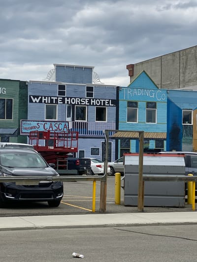 The image shows a street scene in Whitehorse, Canada, featuring the facades of several buildings painted in a colorful, Western-style mural. The central building is labeled "WHITE HORSE HOTEL" in large black letters. To its left, a green building with the partial word "IG" is visible, and to its right, a light blue building is labeled "TRADING CO". Below the "WHITE HORSE HOTEL" sign, a smaller sign reads "OFFICE. INDEPENDENTS ST CASCA". A red hydraulic scissor lift is positioned in front of the "WHITE HORSE HOTEL", with its platform partially raised. Several cars are parked in a lot in front of the buildings. The sky is overcast with clouds. In the foreground, there is a sidewalk and a paved area with yellow bollards and a large grey dumpster. A crumpled paper cup is on the ground in the lower right corner of the image.