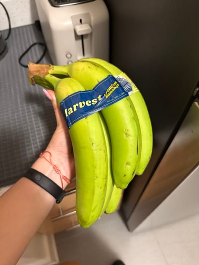 A person's hand, with manicured fingernails, is holding a bunch of four green bananas. The bananas are banded together with a blue label that reads "Harvest Premium Bananas" and "Net wt. 3lbs (1.36k)". The person is wearing a black fitness tracker on their wrist and a red string bracelet. In the blurred background, a white toaster and a grey countertop are visible. The image appears to have been taken indoors, likely in a kitchen, in the city of Yonkers, United States of America.