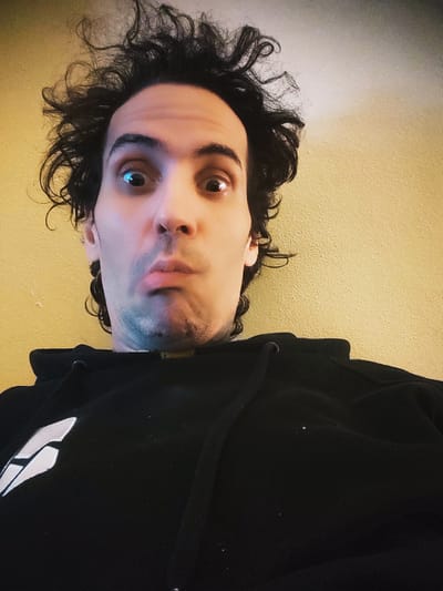 This is a close-up, vertically oriented, low-angle selfie of a person wearing a black hoodie. The person has dark, curly hair and is looking directly at the camera with a neutral expression. Their face is illuminated by a light source from the right, creating highlights and shadows. There is a white logo or design visible on the left side of the hoodie, partially cropped. The background is a textured yellow wall. The lighting suggests it is likely indoors, possibly in the evening or a dimly lit room. There is no visible text in the image.
