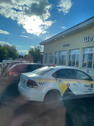 A daytime shot shows a street scene in Bittsa, Russia. Several cars, including a white Yandex taxi with the number 770 on its side and a taxi checker pattern, are parked along the street. A building with signs in Russian for "Shawarma" and "Coffee" is visible. The sky is blue with puffy white clouds. There is sunlight creating glare in the scene.