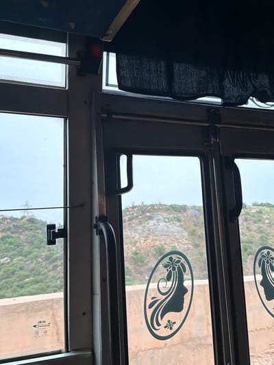 The image shows the interior of a bus, looking out of a window and door. The window on the left has a black handle and a sticker with some text and symbols. The door on the right has a black handle and two oval-shaped decals of a woman's profile with floral designs. Outside the bus, there are hills with greenery and some rocky areas. The sky is overcast. The location is identified as Bethamcherla, India.
