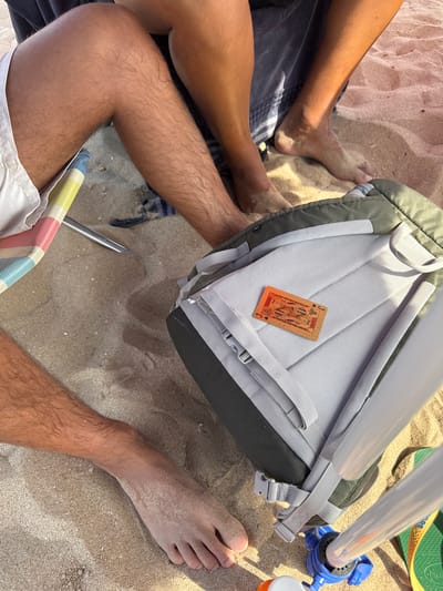 The image shows a close-up view of people's legs and feet on a sandy beach, likely in Costa da Caparica, Portugal, based on the context provided. The sand is light brown and appears somewhat damp in places, with some small shell fragments visible. In the foreground, a person's tanned leg with visible dark hair is seen resting on a striped beach chair, with white shorts visible. To the right, another person's legs and feet, covered in sand, are visible. A backpack, predominantly dark green and grey, is positioned in the center of the frame. A single playing card, featuring an illustration of a man (likely a Jack of Diamonds), is placed on the backpack. The background suggests other people are present, with more legs and sand visible. The lighting indicates it is daytime, with the sun casting shadows and highlighting textures in the sand and on the skin.