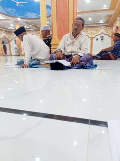 The image depicts an interior scene, likely a place of worship, with a polished white tiled floor. Three men are visible, all seated on the floor on prayer mats. In the foreground, an older man with a mustache and wearing a striped shirt and a sarong is seated and appears to be reading or in prayer. To his left, another man in a white long-sleeved shirt and a sarong is also seated. Further to the right, a third man in a patterned blue shirt and sarong is visible. The background shows architectural elements including columns and decorative wall art, suggesting the location is a mosque. The lighting is bright, indicating overhead illumination. The overall atmosphere is one of quiet contemplation. The specific location of Meunasahmeucat, Indonesia, is not visually apparent from the image itself.
