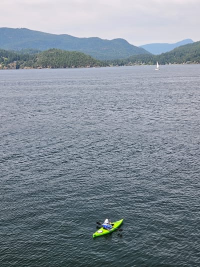 This media file captures an aerial view of a person kayaking on a body of water, likely in West Vancouver, Canada, given the context. The scene is dominated by the dark, rippling surface of the water, which occupies most of the frame. To the left, a lush, tree-covered mountainside slopes down towards the water's edge, with scattered houses visible along the shore. Further in the distance, hazy blue mountains form a layered backdrop against a pale, overcast sky, suggesting a cloudy or misty day.
In the foreground, on the right side of the image, a person is actively paddling a bright green kayak. The kayaker is wearing a blue shirt and a white helmet, and they are holding a black paddle, propelling themselves through the water. The kayak has visible yellow accents. The water around the kayak shows small ripples, indicating movement. In the distance, a solitary white sailboat can be seen near the shore, adding another element to the aquatic scene. The overall impression is one of a peaceful, outdoor activity set against a scenic natural landscape. There is no visible text in the media file.