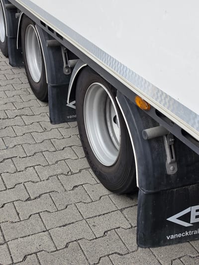 The image shows the side of a white trailer, likely a semi-trailer, with two wheels visible. The trailer has a reflective strip along its lower edge. A black fender is positioned above the larger, front wheel. This fender has a white V-shaped logo and the word "vanecktrain" printed in white lettering on it. The trailer is parked on a paved surface made of interlocking grey concrete blocks. The scene appears to be outdoors, with the lighting suggesting daytime. There are no people visible in the image. The overall impression is of a commercial vehicle parked in a logistics or industrial area.