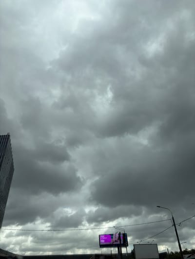 The image captures a cloudy sky over Trekhgorka, Russia, with a modern building on the left and a billboard in the distance. The sky is filled with dark, heavy clouds, suggesting overcast or impending rain. The building is tall and angular, with a reflective facade. In the mid-ground, a billboard with a pink advertisement is visible, featuring an image of clothing. A street light with visible wires is also present in the scene. The bottom portion of the image shows the tops of other structures, possibly buildings or infrastructure, and some greenery. The overall atmosphere is somber due to the cloudy weather.