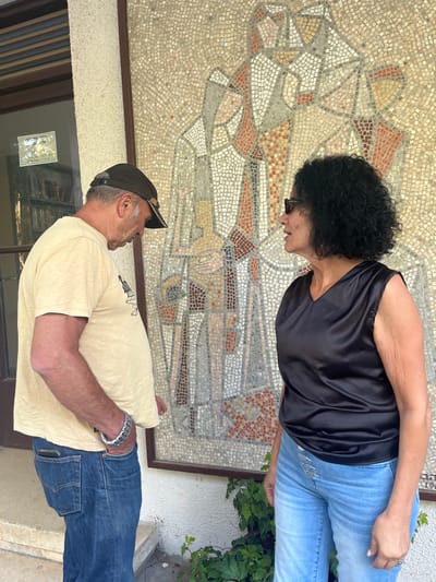 The scene is set outdoors in the Brener Regional Council, Israel, against the exterior wall of a building. Two individuals are present, standing in front of a large, rectangular mosaic artwork.
On the left, a light-skinned male with a dark-colored baseball cap, a light-colored short-sleeved t-shirt, and blue denim jeans stands facing downwards and slightly left. A silver-toned bracelet with a textured design is visible on his left wrist, and a phone is partially visible in his right rear pocket.
On the right, a light-skinned female with dark, curly, shoulder-length hair and dark sunglasses stands. She is wearing a black sleeveless V-neck top, appearing to be satin material, and light blue high-waisted denim jeans featuring a multi-button front closure. She is looking to her left, in the general direction of the male.
The background features a prominent mosaic artwork on a light-colored stucco wall. The mosaic consists of small tiles depicting abstract human-like figures in shades of white, light brown, gray, and reddish-orange, framed by a dark border. To the left of the mosaic, a dark-framed window with internal horizontal blinds shows interior bookshelves filled with books. A small, framed notice is affixed to the window pane. Green leafy plants are visible at the base of the wall, below the mosaic, with a concrete step or ledge immediately in the foreground. The area is illuminated by bright natural daylight.