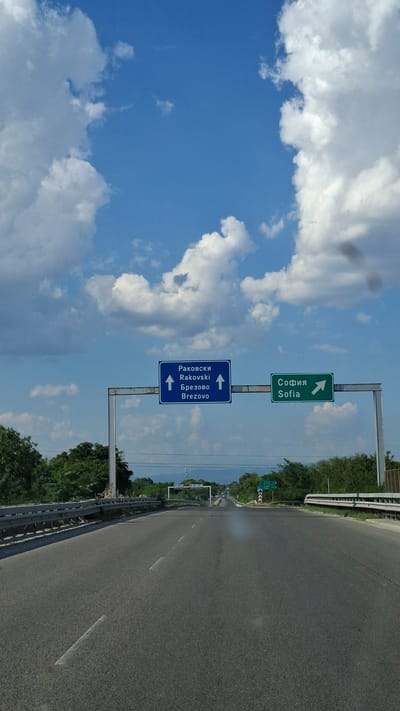 The image depicts a view from a vehicle traveling on a highway in Skutare, Bulgaria. Two blue and green road signs are prominently displayed on an overhead gantry. The blue sign on the left indicates "Rakowski / Rakovski" and "Brezovo / Brezovo" with arrows pointing straight ahead. The green sign on the right indicates "Sofia / Sofia" with an arrow pointing to the right. The highway has multiple lanes and a metal guardrail runs along the right side. Lush green trees line the sides of the road, and mountains are visible in the distance. The sky is a clear blue with scattered white cumulus clouds. The overall scene suggests a typical highway environment with directional signage for nearby towns and the capital city.