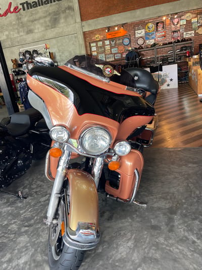 This image captures the interior of a motorcycle-themed establishment located in Ban Yid, Thailand. A large, two-toned touring motorcycle, identified by a "RIDE COMMANDER THAILAND" emblem on its fairing, dominates the foreground. The motorcycle features a peach-bronze and black paint scheme, with the peach-bronze covering the front fender, fairing, and saddlebag components, and a black section on the upper fairing and fuel tank. It is equipped with dual chrome headlights, an amber front turn signal, and various chrome accents on the handlebars, engine guard, and front forks. The motorcycle is parked on a mottled gray polished concrete floor.

To the left and slightly behind the primary motorcycle, a second, predominantly black motorcycle is partially visible. The background wall on the left is a light gray concrete surface adorned with large red, three-dimensional letters spelling "Ride Thailand." Below this, a white grid display rack holds multiple helmets or headwear. The wall on the right features an exposed red brick pattern, heavily decorated with numerous vintage-style metal signs, many depicting motorcycles, road themes (including a "HARLEY-DAVIDSON STREET" arrow sign and a "ROUTE 66" shield), and garage motifs. Below these signs, multi-tiered metal shelving units display folded apparel and hats. The floor on the right side of the establishment transitions to a polished wooden surface with a distinct diagonal stripe pattern. No individuals are present within the scene.