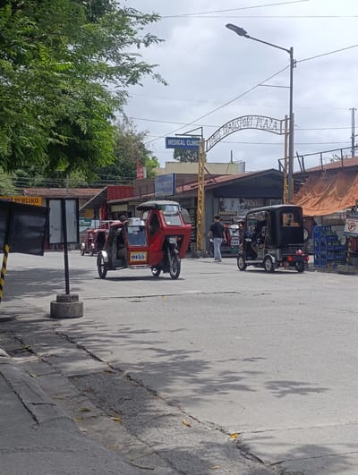This is a street scene in Imus, Philippines, under a cloudy sky. In the foreground, a red and silver motorized tricycle with the number 0155 on its side is parked. To the right, a black motorized tricycle is also parked. Behind them, other tricycles and a few pedestrians are visible. Several buildings line the street, including one with a sign that reads "MEDICAL CLINIC." An arched sign overhead displays "IMUS TRANSPORT PLAZA." The street is paved and shows some wear and tear, with cracks and patches. Trees and shadows from foliage are present on the road.
