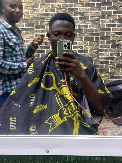 The image captures a mirror selfie taken by a person in what appears to be a barbershop or hair salon. The main subject, a young person with dark skin and short, dark hair, is positioned centrally, looking directly into the camera lens of a smartphone. They are wearing a black barbershop cape adorned with yellow illustrations of barber tools, including large scissors, combs, and what resemble clippers or battery symbols. The word "BARBER" is also visible in yellow text on the cape, appearing mirrored in some instances. The smartphone, an iPhone with a distinctive triple-camera system and a light green protective case, is held up by the subject's right hand, covering part of their lower face.
The background behind the subject, as reflected in the mirror, consists of a textured wall with a brown and tan brick pattern. Below the main subject in the mirror's reflection, another individual is partially visible. This second person, likely a barber or another customer, is wearing a plaid shirt with shades of blue, green, and white. Only their upper torso and head are seen, with their gaze directed slightly away from the primary subject.
A thin, bright red cord, possibly from headphones or a charging cable, is draped over the black cape, extending from the upper right of the frame towards the left. On the far right edge of the mirror, a white frame is visible, beyond which appears to be a patch of artificial green grass or carpet. The overall lighting suggests an indoor setting, likely under artificial light.