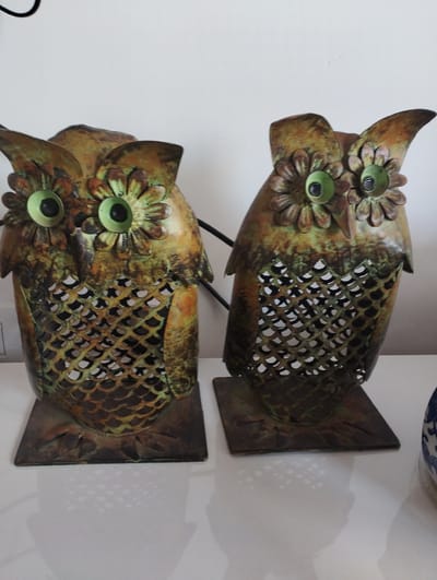 The image shows two decorative metal owl figurines displayed on a white surface, likely a shelf or table. The owls are a rustic bronze and green color, with large, round, bright green eyes. Their bodies are hollow and feature a cut-out pattern resembling scales or diamond shapes, allowing light to pass through. Each owl stands on a square metal base. The background is a plain white wall. The lighting appears to be neutral, suggesting it might be daytime indoors, or a well-lit room. No people, text, or specific time or weather cues are visible. The scene is domestic or retail, showcasing decorative items.