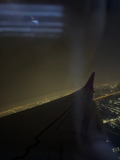 An aerial view at night captures a portion of an aircraft's dark grey wing, prominently positioned in the middle ground, extending from the lower left towards the upper right. The wing displays faint markings, including "D-HHK," and features a small section of a purple or dark red wingtip. Below the wing and across the horizon, the urban sprawl of Racha Thewa Subdistrict, Thailand, is illuminated by a dense grid of warm-toned city lights. The sky above is dark and hazy, with a greenish-brown tint visible near the horizon. A distinct, bright vertical beam of light, originating from an unseen source, cuts through the atmospheric haze in the upper-middle section of the frame. Reflections of interior cabin lights or structural elements are present as faint horizontal streaks and a small luminous point in the upper left corner of the image.