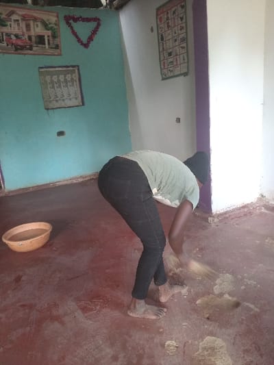 A person in a gray shirt, black pants, and a black head covering sweeps debris from a dirt floor. The person is barefoot. A small, round, orange bowl sits nearby on the floor. The walls are painted blue and white and decorated with artwork. A purple strip is present at the top of the wall. The floor has uneven areas of brown and tan dirt, with some sections appearing swept.