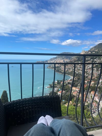 The image shows a person sitting on a balcony overlooking the Mediterranean Sea. The person is wearing blue jeans and white socks. They are sitting on a wicker chair, facing the viewer. The balcony is lined with a black metal railing. The scene is taken from the town of Roquebrune-Cap-Martin in France, located at latitude: 43.763, longitude: 7.464.  The blue sea stretches out in front of them, while the town is situated on a hillside in the distance, with buildings and trees. The sky is blue with white clouds. There is a small boat in the distance, navigating the sea. The scene appears peaceful and serene, with a beautiful view.