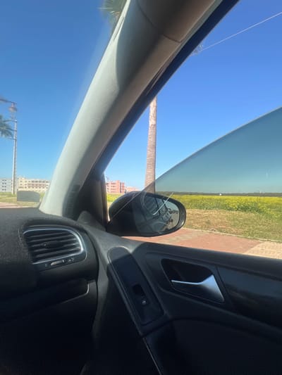 The image depicts an exterior scene from the perspective of a vehicle's front passenger seat in Beni Mellal, Morocco, under a clear, bright blue sky. The vehicle's interior components visible include a grey A-pillar on the left, a black dashboard section with a chrome-trimmed air vent on the lower left, and a black door panel on the lower right featuring a silver door handle and a window control switch.
The passenger side view reveals a cracked exterior side mirror, showing multiple fracture lines across its reflective surface. A thin, light-colored string or wire passes diagonally through the frame, stretching from the upper left portion of the window opening across the side mirror.
Beyond the vehicle, a paved roadside or sidewalk runs adjacent to a large, expansive field covered in bright yellow flowering plants. In the midground, a tall, bare palm tree trunk stands prominent, with additional palm trees visible on the far left, partially obscuring a streetlight. The background features several multi-story buildings; some on the left appear to be completed structures, while others further in the distance, particularly to the right, exhibit signs of ongoing construction with exposed brickwork. The overall environment suggests an urban periphery meeting agricultural land.