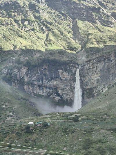 The image depicts a majestic waterfall cascading down a sheer cliff face in the mountainous terrain of Yamling, India. The waterfall is the central focus, with its white water creating a dramatic contrast against the rugged, grey rock of the cliff. The surrounding landscape is characterized by steep, green-covered slopes that appear terraced in places, suggesting agricultural activity. A small cluster of buildings, including a white tent and a house with a green roof, is visible on one of the lower slopes, nestled amongst the vegetation. A fine mist generated by the waterfall hangs in the air near the base of the falls and on the surrounding slopes. The sky above is overcast, casting a diffused light over the scene. The overall impression is one of natural grandeur and remote, rustic habitation.