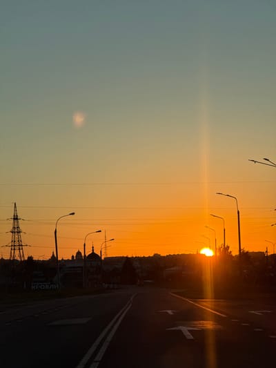 The image shows a sunset over a highway in Morózovka, Russia. The sun is setting behind a silhouette of buildings and trees, casting a warm orange glow across the sky. The sky transitions from a soft blue at the top to a vibrant orange near the horizon. In the foreground, the highway is dark with visible lane markings. Several streetlights line the right side of the road, their silhouettes stark against the bright sky. A large power pylon is visible on the left side of the image. The overall scene depicts a serene and picturesque view of a sunset in a semi-urban environment.