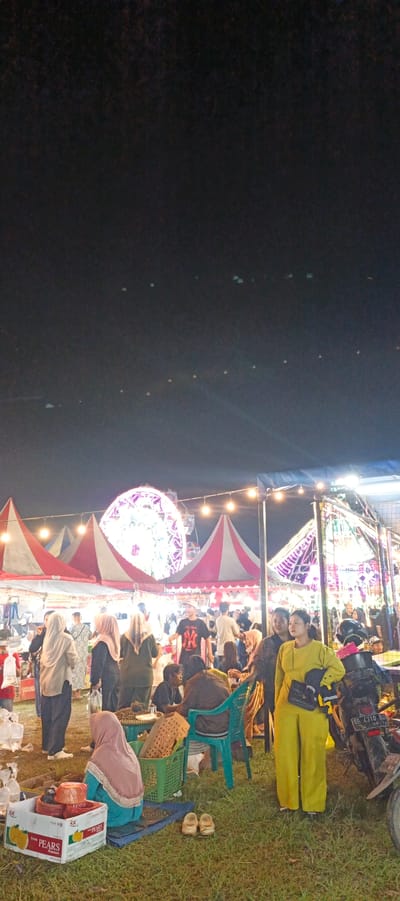 This image captures a lively night scene at a fairground in Banding Agung, Indonesia. A Ferris wheel, illuminated with purple lights, stands prominently in the background, suggesting a festive atmosphere. Numerous tents with red and white awnings are set up, likely housing stalls or attractions. A crowd of people, many wearing traditional attire, are milling about, engaged in various activities. Some are gathered around stalls, while others are seated, observing the surroundings. A woman in a bright yellow outfit is standing near a parked motorcycle, holding a bag. In the foreground, a box labeled "PEARS" and some footwear are visible on the grassy ground. The overall impression is one of a bustling community event under a dark, star-studded sky.