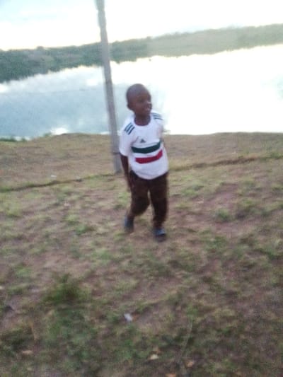 A blurry, grainy photograph captures a single young male child standing outdoors in Central Kasipul ward, Kenya. The child, appearing to be of early primary school age, has dark skin and short, dark hair. He is wearing a white t-shirt with a distinctive horizontal stripe pattern across the chest consisting of a green stripe, followed by a white stripe, and then a red stripe. He also wears dark brown trousers and dark closed-toe footwear. The child is positioned slightly off-center to the left, facing generally towards the camera but with his head turned slightly to his right, displaying a subtle upward curve at the corners of his mouth. Behind the child and slightly to his left, a tall, slender, light-colored pole extends vertically out of frame at the top. The ground in the foreground and midground is uneven, covered with sparse brown and green vegetation interspersed with exposed dirt. In the background, beyond the immediate land, a calm body of water, likely a lake or wide river, reflects the overcast sky. A faint, distant landmass with vegetation is discernible across the water. The overall lighting is soft, consistent with an overcast day. No other individuals or interactions are visible.
