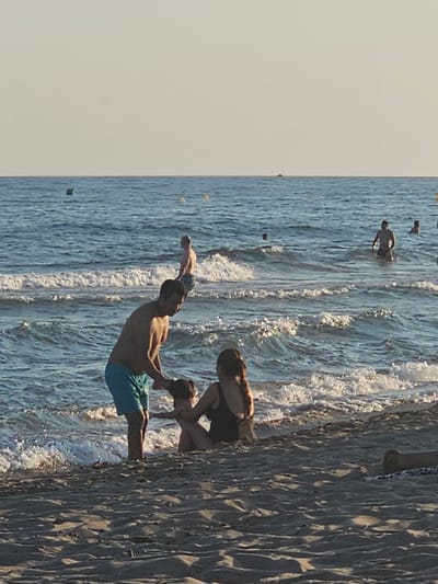 The media file captures a scene at the beach in Castelldefels, Spain. The image shows several people in the water and on the sand. In the water, a group of individuals are swimming and playing, with some appearing to be diving or jumping. The water is a vibrant blue with white foamy waves breaking near the shore. The sand on the right side of the image is a light brown color and shows textured patterns from the waves. The sky is a pale, hazy color, suggesting it might be late afternoon or early morning, or possibly a slightly overcast day. The overall atmosphere appears to be one of leisure and enjoyment, typical of a beach outing. There is no visible text in the image.