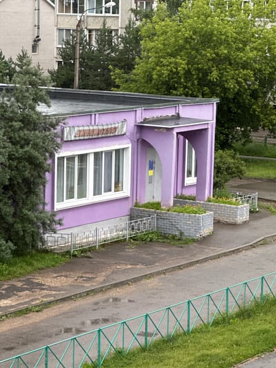 The image shows a small, vibrant purple building with arched doorways and multiple windows. The building appears to be part of a larger complex, with adjacent structures and greenery visible. A paved walkway runs alongside the purple building, bordered by a bright green metal fence and a patch of grass. Two brick planters with small yellow flowers are situated next to the entrance of the purple building, adding a touch of color to the scene.
The perspective suggests the photo was taken from an elevated position, possibly from an upper floor of a nearby building or a drone. The lighting indicates it is daytime, and the lush greenery implies a warm weather season, likely spring or summer. The overall setting appears to be a residential or public area within a Russian town, given the location context provided as Rzhev, Russia. There are no people or animals visible in the frame.
Notable details include the slightly weathered appearance of the pavement and the presence of a utility pole with wires extending from it. A narrow, elongated window with some form of writing or signage on it is visible on the side of the purple building. The architecture suggests a functional, possibly institutional or commercial, use for the purple structure. The scene is static, with no activity taking place, presenting a snapshot of the environment.