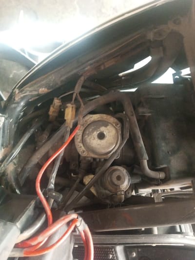 A close-up shows the engine compartment of a scooter or motorcycle. Visible components include the carburetor, fuel lines, wiring harnesses, and electrical connectors. The carburetor appears worn, with a dark coating on the surface. Hoses and wires are routed around the engine components. The overall scene is dimly lit, and the compartment appears confined.