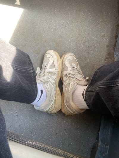 The image shows a person sitting, with their feet resting on the floor. They are wearing dark denim jeans and off-white athletic shoes with white socks that have a black Nike swoosh. The floor appears to be gray and speckled, possibly a public transportation setting like a bus or train. There is no information to support the location in Matyashivka, Ukraine.