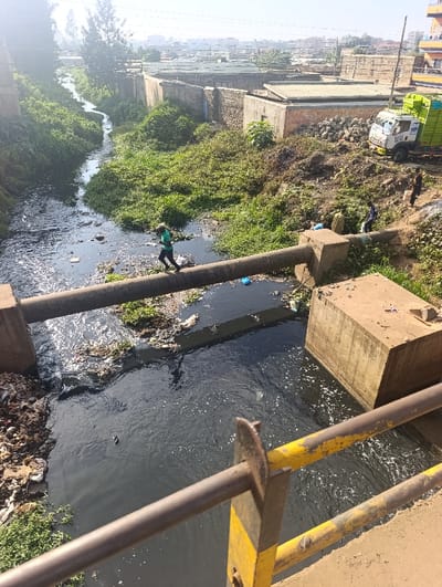 A polluted river flows through a densely populated area. Debris lines the banks and floats in the dark water. A person walks across a large pipe spanning the river. Nearby, other figures stand near a garbage truck. Buildings and vegetation border the river. The view is framed by a yellow-striped railing.