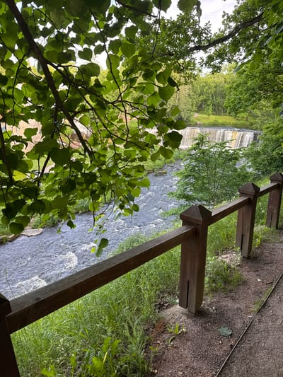 The image depicts a scenic view of the Keila-Joa waterfall and the Keila River in Keila-Joa alevik, Estonia. In the foreground, a wooden fence with dark brown posts and rails runs diagonally across the scene, separating a gravel path from a grassy embankment. Lush green foliage from tree branches overhangs the upper left portion of the image. The Keila River flows rapidly through the mid-ground, with white water indicating a fast current. Beyond the river, the Keila-Joa waterfall cascades over a rock formation. The background consists of dense green trees and a hint of a building, possibly the Keila-Joa Manor, on the left. The sky is overcast, suggesting a cloudy day. No people are visible in the image.